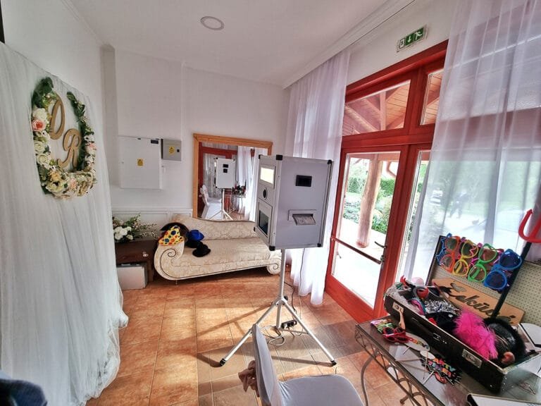 Fotóbox bérlés. Elegáns beltéri helyszínen elhelyezett fotóbox, amely mellett egy kanapé és különböző mókás kiegészítők várják a vendégeket. A háttér romantikus esküvői dekorációval díszített, a fotóautomata pedig azonnali fényképek nyomtatására is képes. Tökéletes választás esküvőkre, céges rendezvényekre, családi eseményekre és születésnapokra, hogy maradandó élményeket teremtsen!
